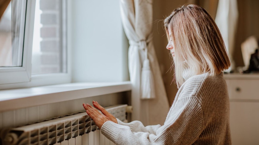 Consumer nz website promo image young blond woman in long winter beige sweater is warming herself near a heater width
