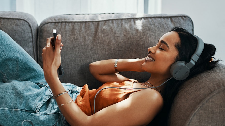 Shot of a young woman using a smartphone and headphones on the sofa at home