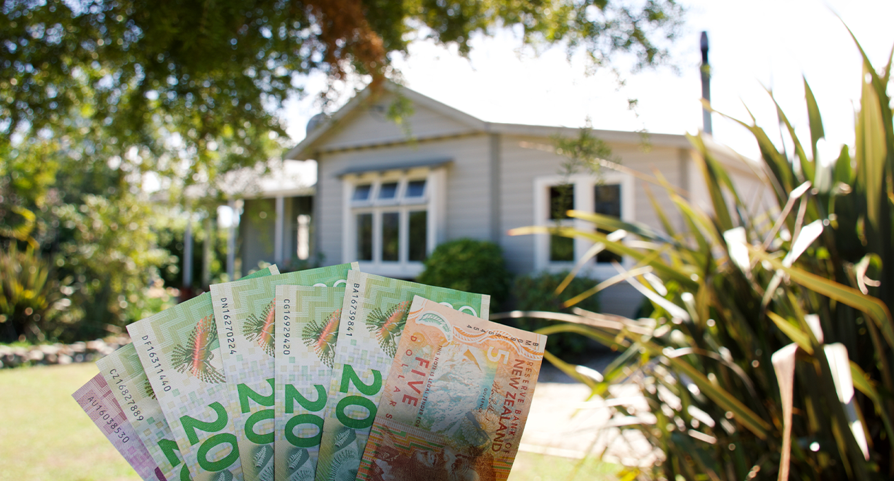 Hand holding up New Zealand money with a house out of focus in the background.